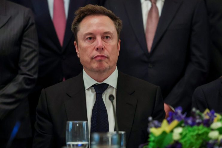 Elon Musk in a dark suit and blue tie, seated at a table with a glass and microphone.