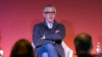 Man with grey hair and glasses speaking, wearing a grey jacket and blue jeans, sitting in a chair with arms crossed.