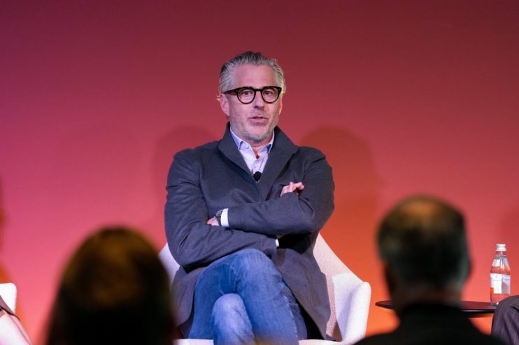 Man with grey hair and glasses speaking, wearing a grey jacket and blue jeans, sitting in a chair with arms crossed.