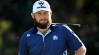 Bearded man in white Topgolf hat and blue patterned polo shirt, golf club on shoulder.