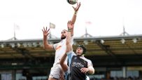 Rugby players leaping for a lineout ball during a match, stadium crowd in background