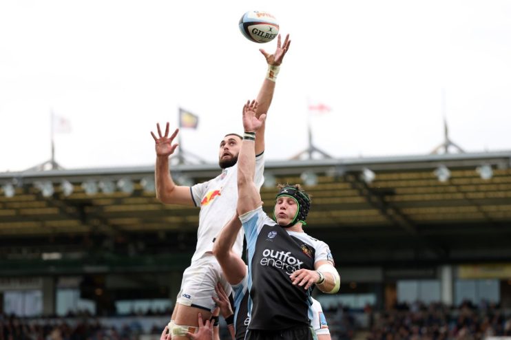 Rugby players leaping for a lineout ball during a match, stadium crowd in background