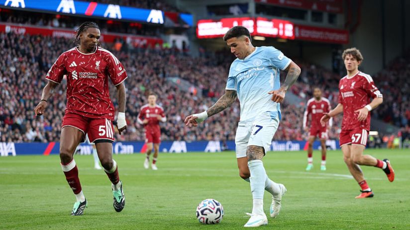 Two soccer players in red and light blue jerseys compete for the ball on a green field during a match.