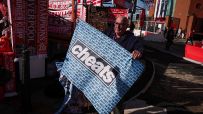 Man holding a cheats banner with GUILTY 114 text, Liverpool FC merchandise in background