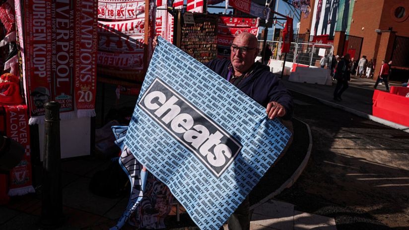 Man holding a cheats banner with GUILTY 114 text, Liverpool FC merchandise in background