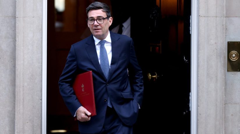 Andy Burnham, Mayor of Greater Manchester, in a dark suit holding a red folder, exiting a doorway.
