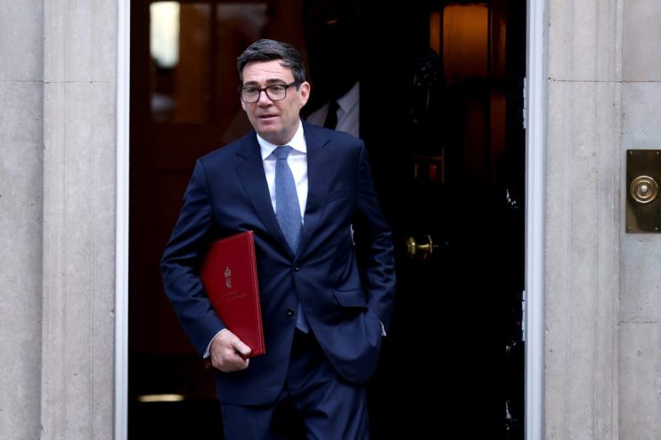 Andy Burnham, Mayor of Greater Manchester, in a dark suit holding a red folder, exiting a doorway.