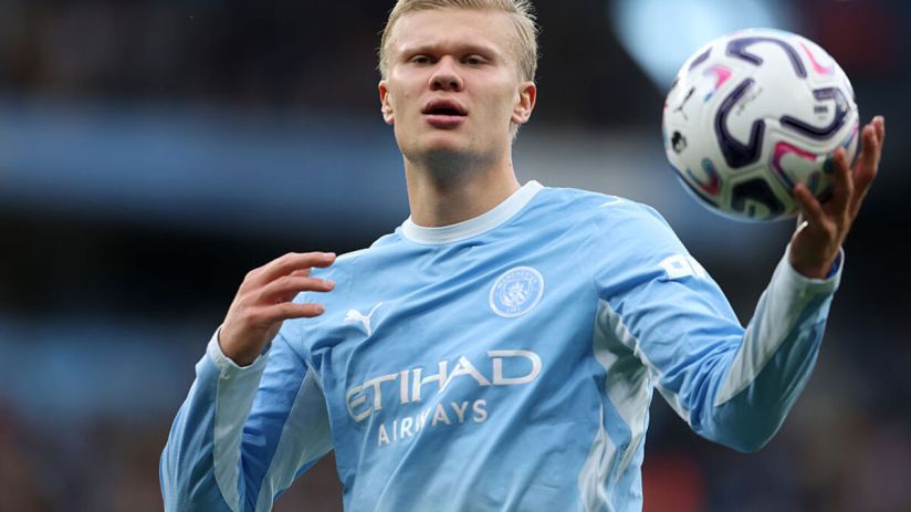 Erling Haaland in Manchester City light blue jersey holding a football, looking forward on the field