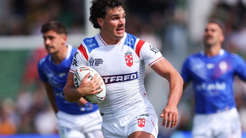 A male rugby player in a white England jersey runs with the ball on a sunny day.