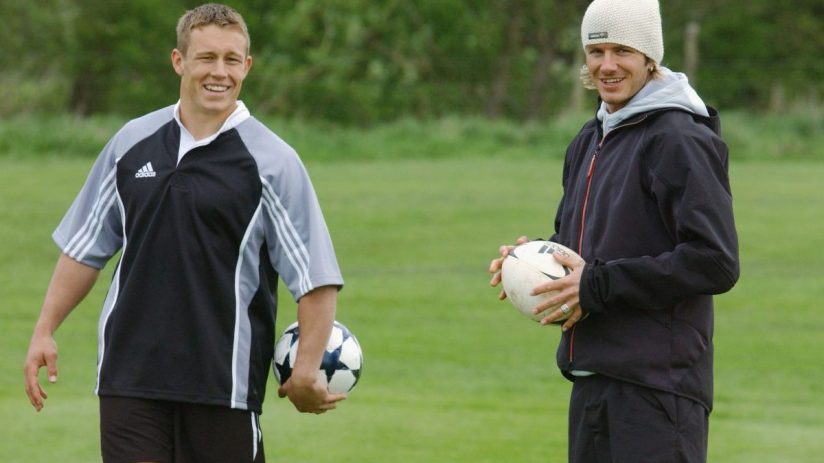 Jonny Wilkinson and David Beckham holding footballs on a grassy field, smiling.