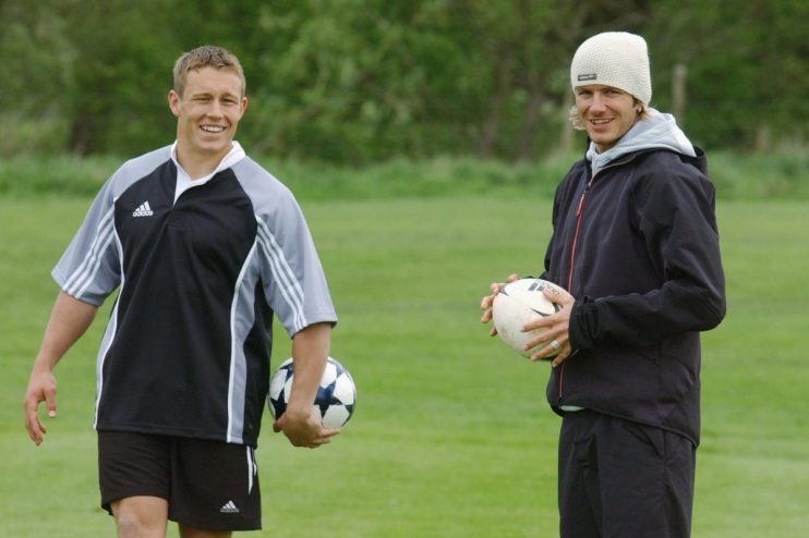 Jonny Wilkinson and David Beckham holding footballs on a grassy field, smiling.