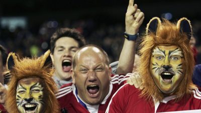 Excited sports fans in red shirts, some with tiger face paint and lion wigs, cheering with a banner