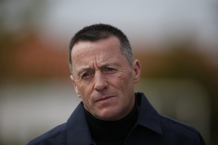 A man with short dark hair and blue eyes, wearing a dark blue jacket over a black turtleneck, looking intently.