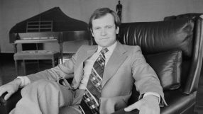 Man in suit and patterned tie sitting in a leather armchair, an Oscar-like statue and piano in background.