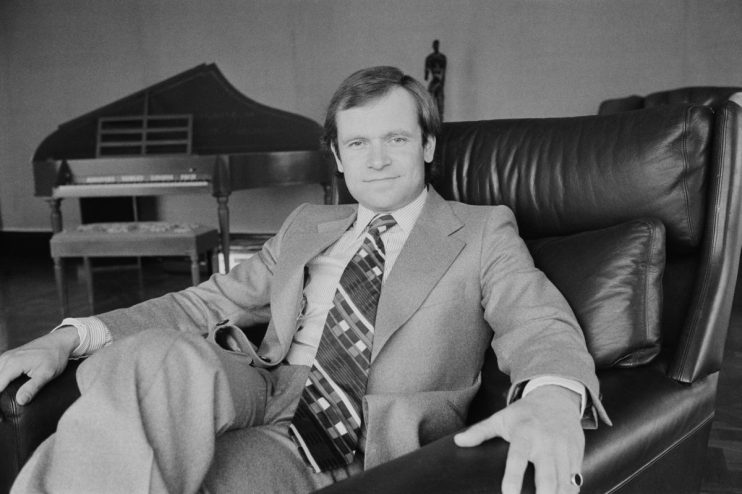 Man in suit and patterned tie sitting in a leather armchair, an Oscar-like statue and piano in background.