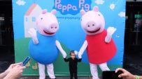 A young child stands between two Peppa Pig characters, with adults photographing them.