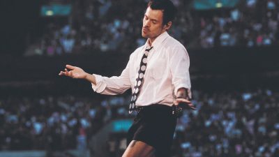 Harry Styles in white shirt, black shorts, and polka dot tie on stage, performing for a crowd.