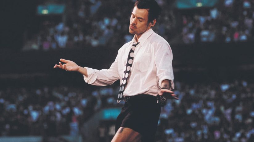 Harry Styles in white shirt, black shorts, and polka dot tie on stage, performing for a crowd.