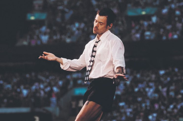 Harry Styles in white shirt, black shorts, and polka dot tie on stage, performing for a crowd.