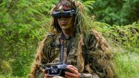 Soldier in ghillie suit and VR headset operating a drone remote control for defence startups.