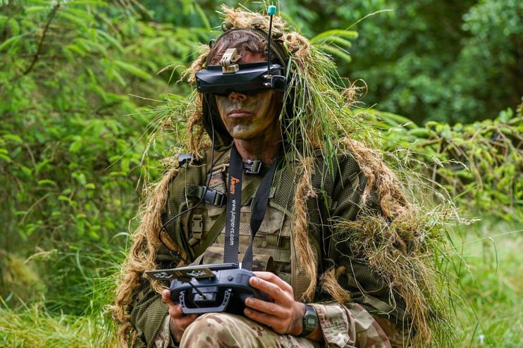 Soldier in ghillie suit and VR headset operating a drone remote control for defence startups.