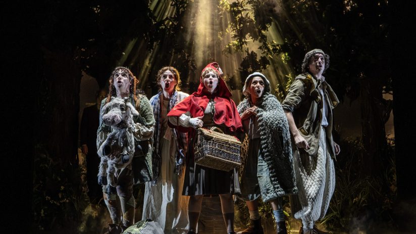Cast of Into the Woods London looking up in fear, light beams through the dark forest canopy.
