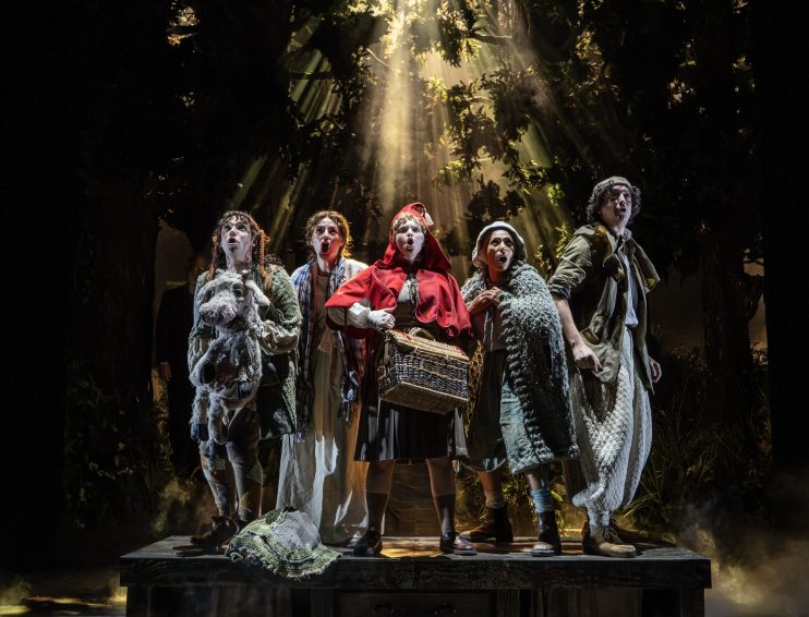 Cast of Into the Woods London looking up in fear, light beams through the dark forest canopy.