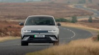 Gray Ioniq 5 electric car driving on a winding road through a dry, grassy landscape with a driver visible.