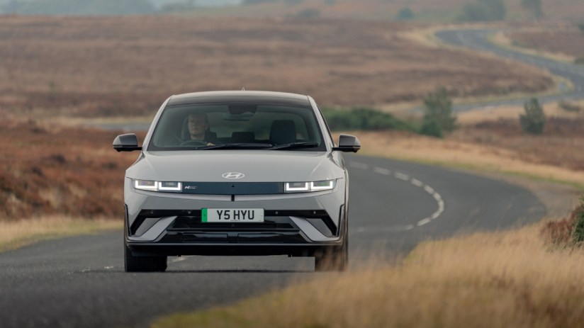 Gray Ioniq 5 electric car driving on a winding road through a dry, grassy landscape with a driver visible.