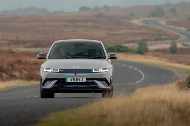 Gray Ioniq 5 electric car driving on a winding road through a dry, grassy landscape with a driver visible.