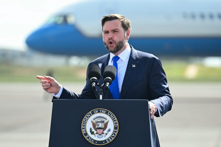 JD Vance speaking at a podium with the Vice Presidents seal, pointing, with a plane in the background.