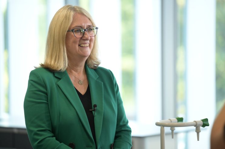 Jacqui Smith, former Home Secretary, smiling in a green blazer with a microphone clipped on, indoors.