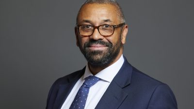 James Cleverly, a man with a beard and glasses, smiling in a navy blue suit and spotted tie