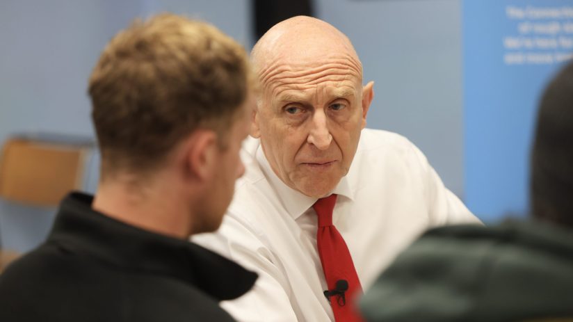 John Healey, bald man in white shirt and red tie, listening intently to someone off-camera.