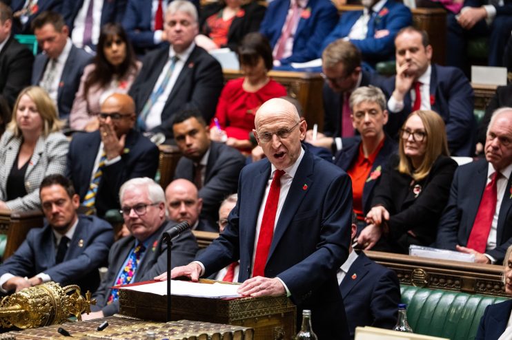 John Healey delivering the Budget speech in Parliament, surrounded by MPs.