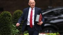 John Healey, a bald man in a dark suit and red patterned tie, walks holding red and green folders.