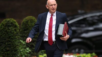 John Healey, a bald man in a dark suit and red patterned tie, walks holding red and green folders.
