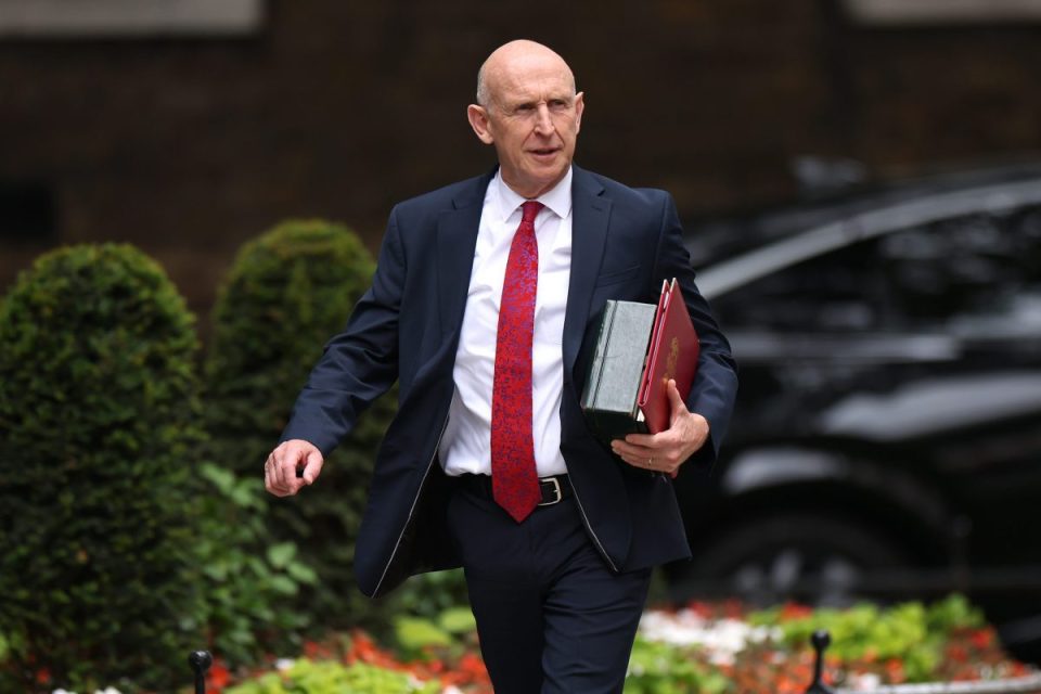 John Healey, a bald man in a dark suit and red patterned tie, walks holding red and green folders.