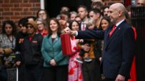 John Healey, in a suit, holds a red budget box aloft before a crowd outside a building.