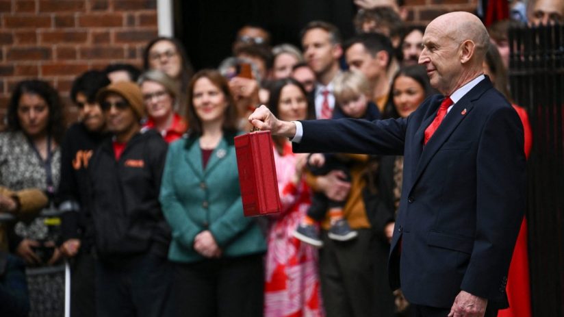John Healey, in a suit, holds a red budget box aloft before a crowd outside a building.