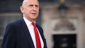 John Healey, bald, in a navy suit and red tie, stands outside a building, likely Number 10 Downing Street.