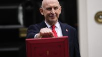 John Healey, MP, smiling and holding the red Budget box outside a building