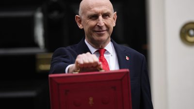 John Healey, MP, smiling and holding the red Budget box outside a building