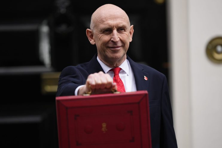 John Healey, MP, smiling and holding the red Budget box outside a building