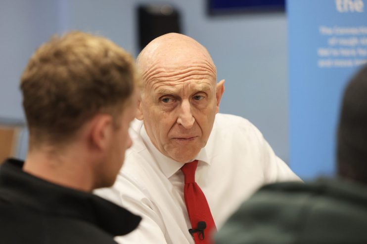 John Healey, bald man in white shirt and red tie, listening intently to someone off-camera, facing scrutiny.