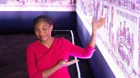 Kemi Badenoch in a red dress gesturing towards a tapestry-like display with historical scenes and text THE WELFARE PARTY