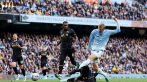 Manchester City player Erling Haaland in action during a football match, with other players and fans in the background.