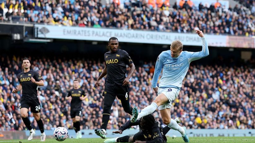 Manchester City player Erling Haaland in action during a football match, with other players and fans in the background.