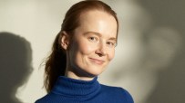 Marit Rodevand smiling, wearing a blue turtleneck, with red hair tied back, against a textured background.