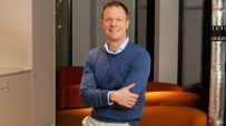 Mark Fairless, smiling, in a blue sweater and beige pants, arms crossed, in a modern office.